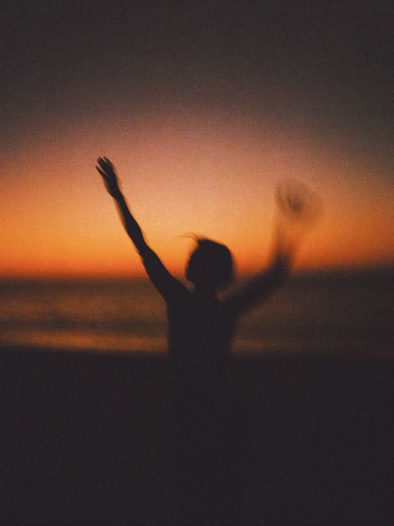 A dancer silhouetted against a deep orange sunset over the water, arms sweeping upward in a blur of motion